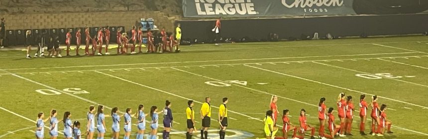 Soccer players from opposing teams, one team in blue and the other in orange, lined up on a field