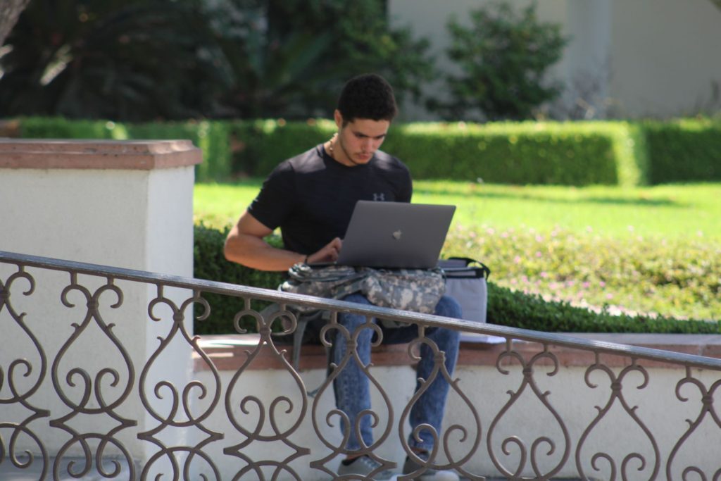 One student working on a laptop, sitting on a ledge outdoors