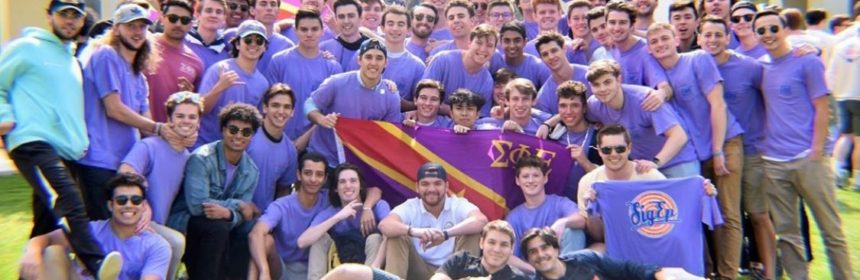 Group of male students, all wearing purple shirts, holding fraternity banner