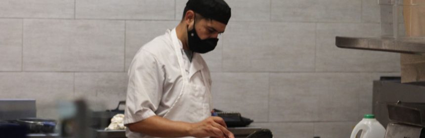 Chef wearing white coat and black hat cooks vegetables in a pan behind a counter