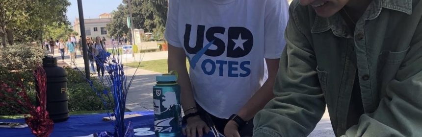 Two people wearing a shirt saying "USD votes" smiling down with a pen and signing a piece of paper