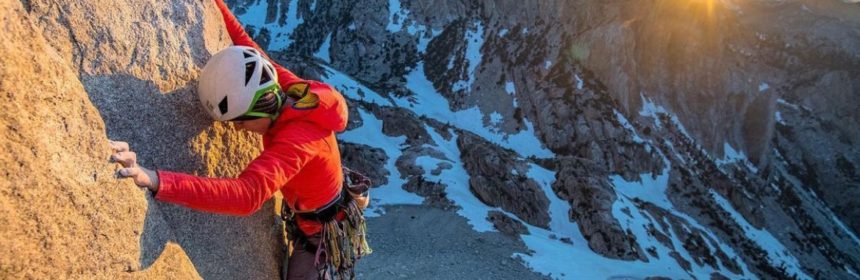 person in red jacket and a helmet on the side of a cliff with the sun peeking over a mountain in the back
