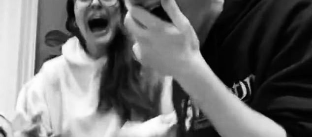 black and white screenshot of boy smiling and covering his mouth with people celebrating in the back
