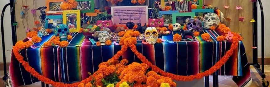 table with orange flowers in front and colored paper decorations behind it. differently colored frames with peopels faces and skulls on table